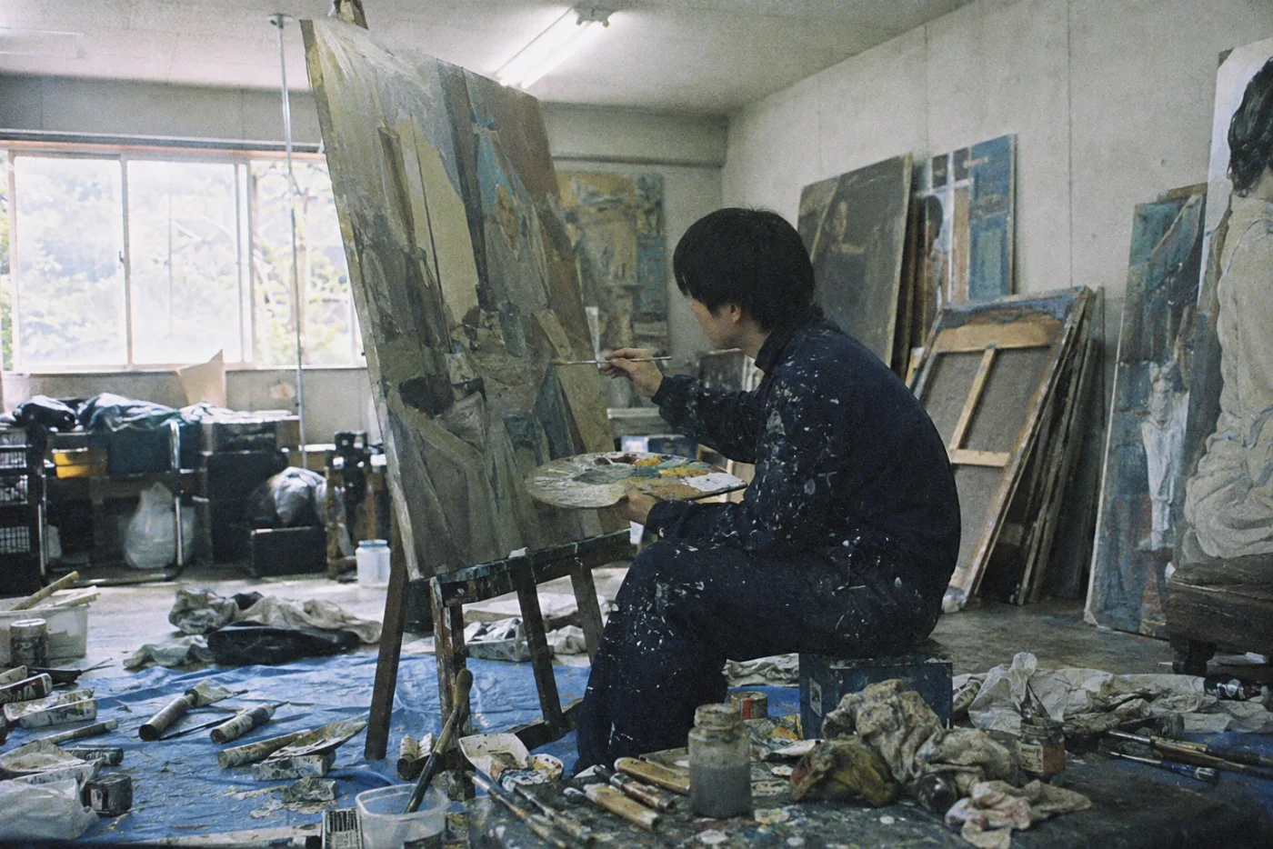 美術大学のアトリエで、描きかけの油絵に向かう学生。床には絵の具のチューブや汚れた布が散らばっている