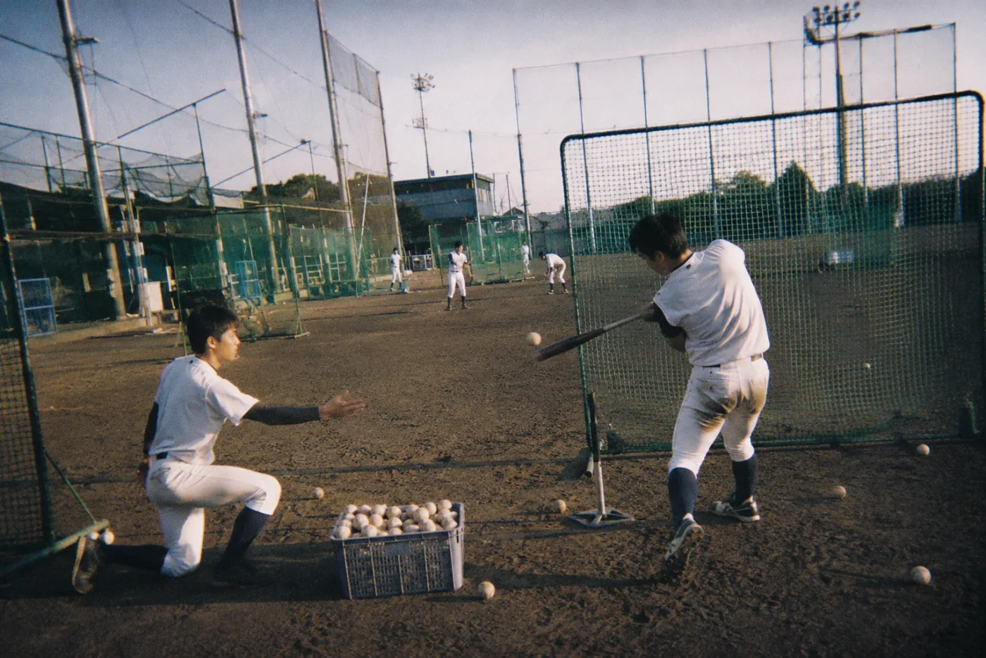 夕方のグラウンドでティーバッティングを繰り返す野球選手。足元にはボールの入った籠がある